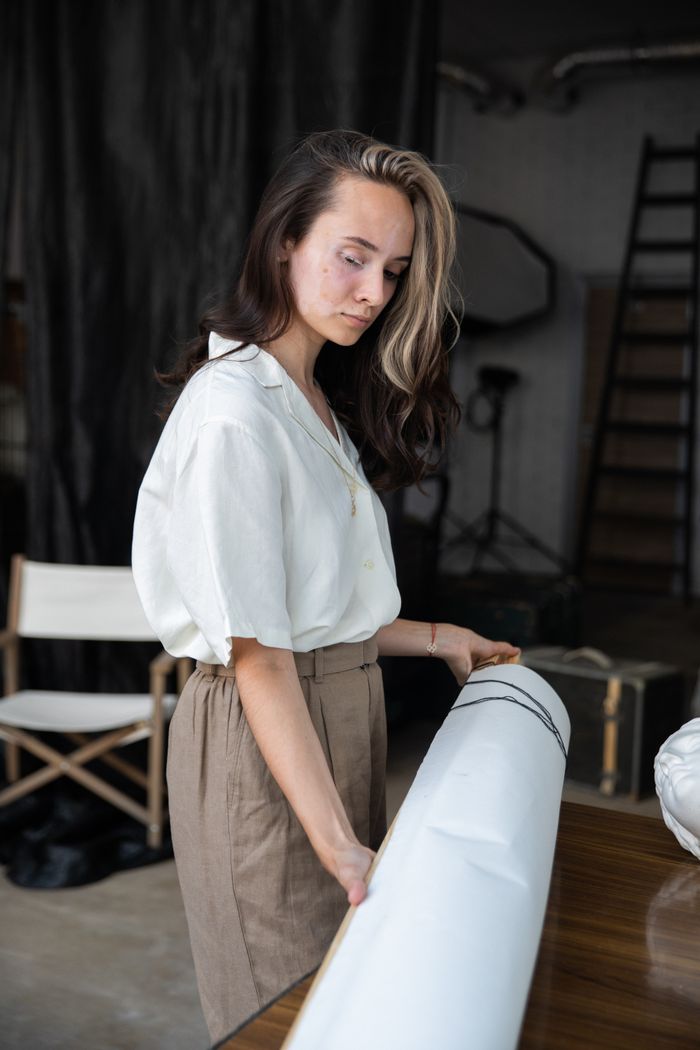 A woman with long hair stands indoors, holding a rolled paper in a stylish setting.